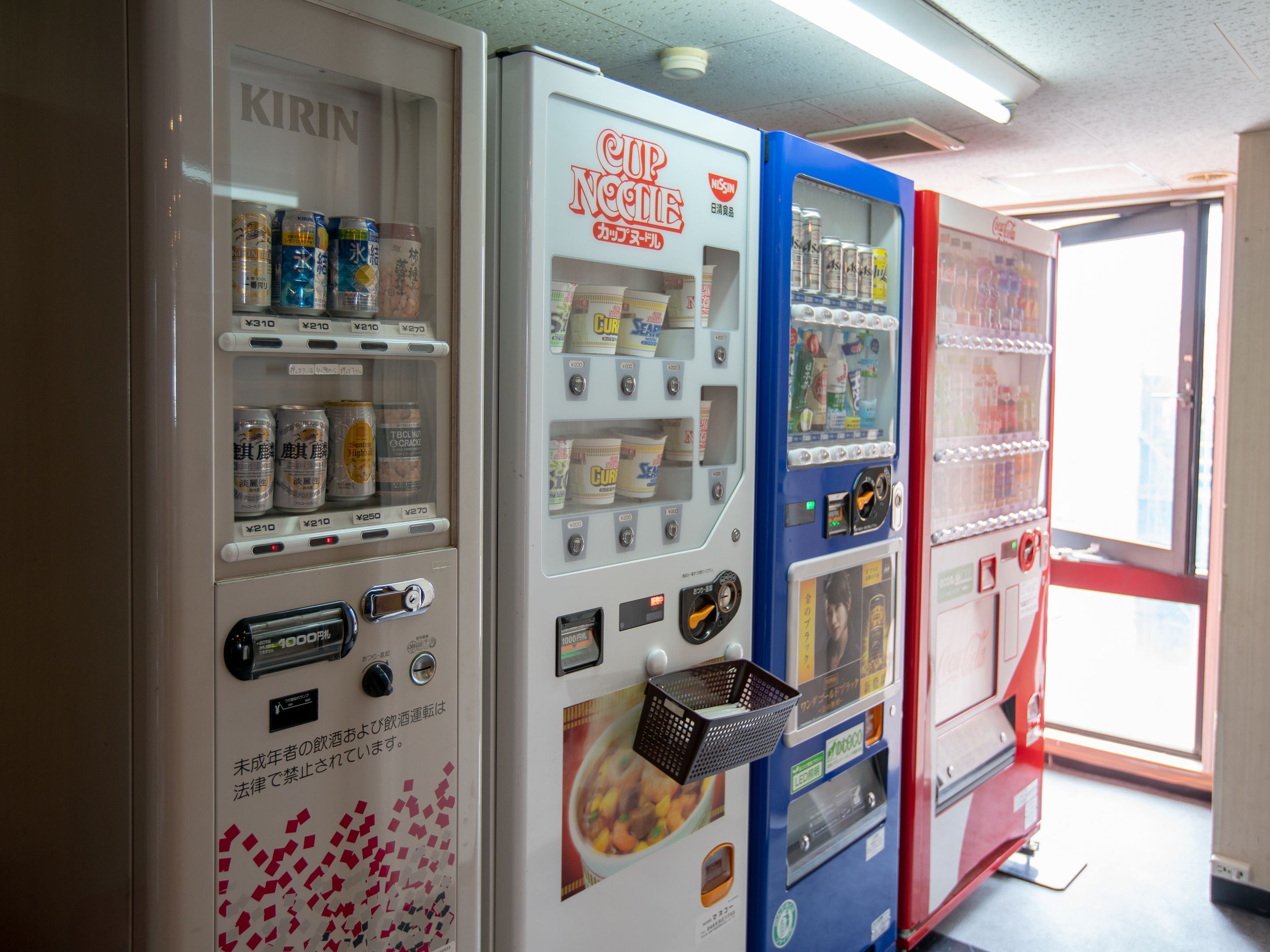 Vending machines with various beverages and snacks, a red door, and a window with natural light.