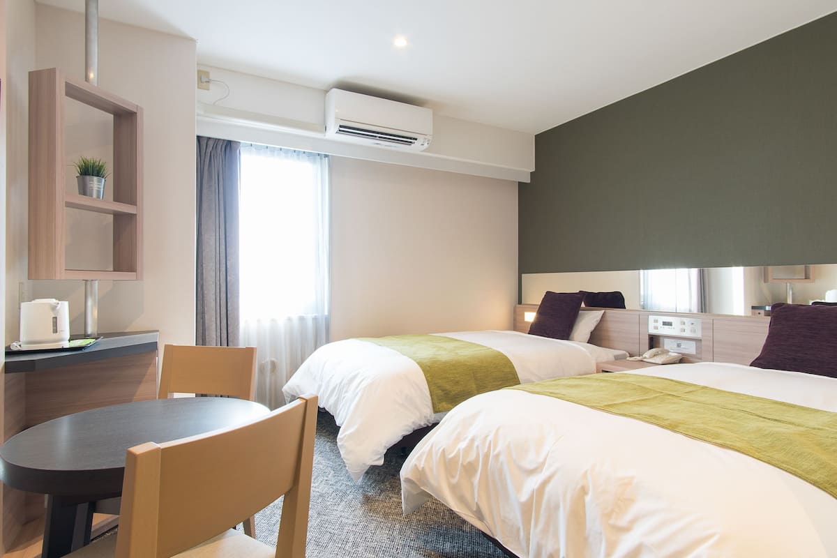 A hotel room with two beds, a desk with a chair, a small table, and a window with curtains.