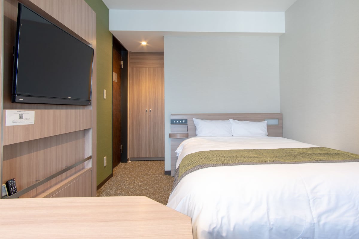 A hotel room with a bed, a TV mounted on the wall, a wooden wardrobe, and a small desk with a chair.