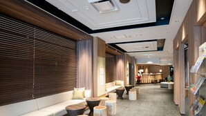 A modern hotel lobby with a long reception desk, seating area with sofas and round wooden tables, and a counter with a menu board. - Fujinomiya Fujikyu Hotel (Fujinomiya)