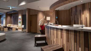 A modern reception area with a wooden reception desk, seating area with sofas and round tables, and a wall-mounted security camera. - Fujinomiya Fujikyu Hotel (Fujinomiya)