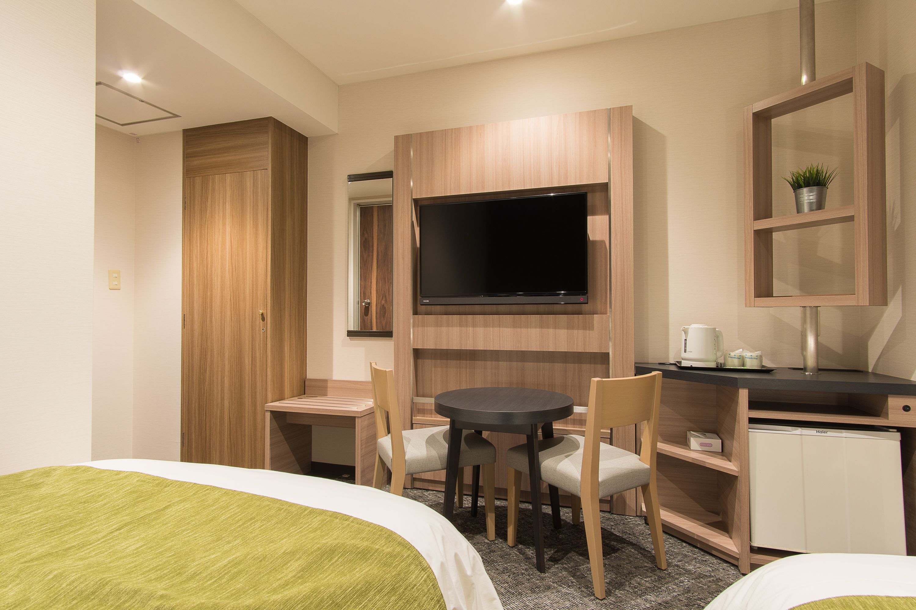 A hotel room with a bed, a desk with a chair, a television, a small cabinet, and a shelf with a plant.
