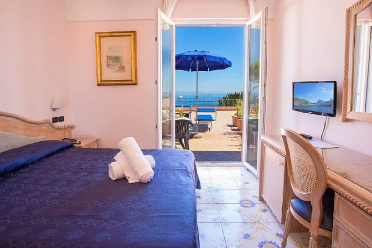 Un dormitorio con cama, escritorio, silla, televisor y balcón con vista al mar.