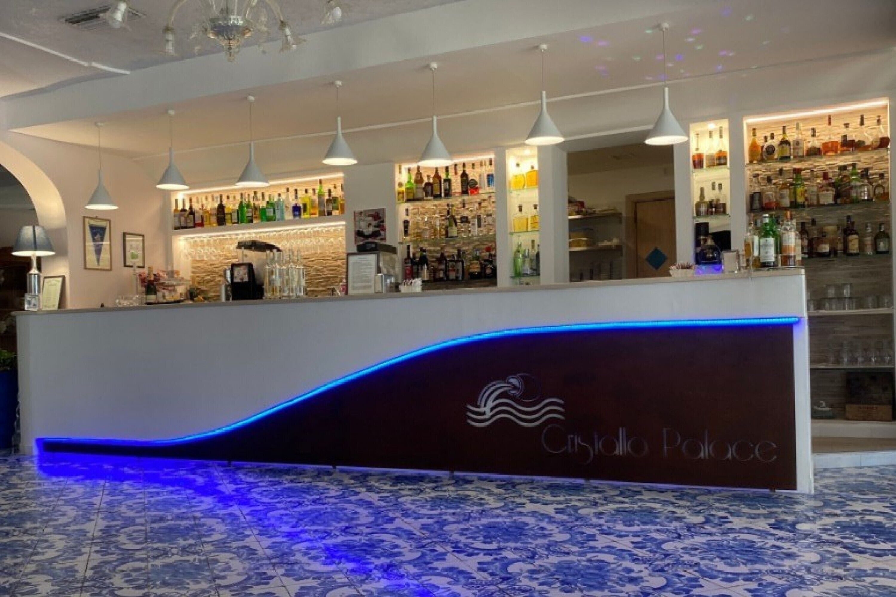 Zona de bar con una recepción de diseño curvo, lámparas colgantes y una variedad de botellas y vasos en estanterías.