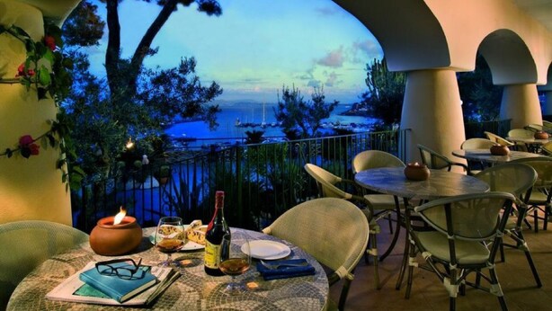 A table set with a bottle, glasses, and a book, overlooking a body of water with boats. - Hotel Terme Cristallo Palace  (Casamicciola Terme)