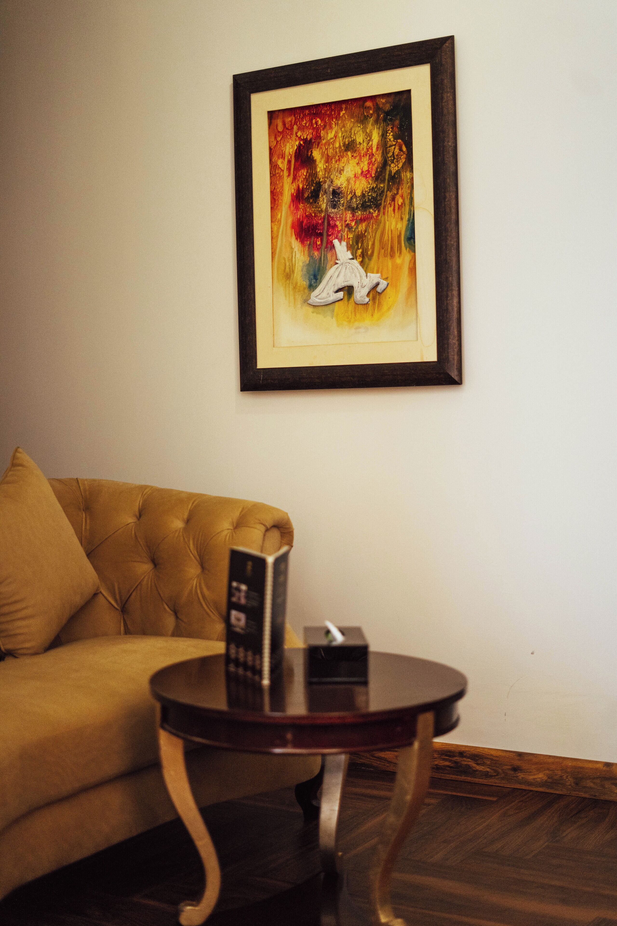 A room with a tufted sofa, a small round table with a remote control, and a framed abstract painting on the wall.