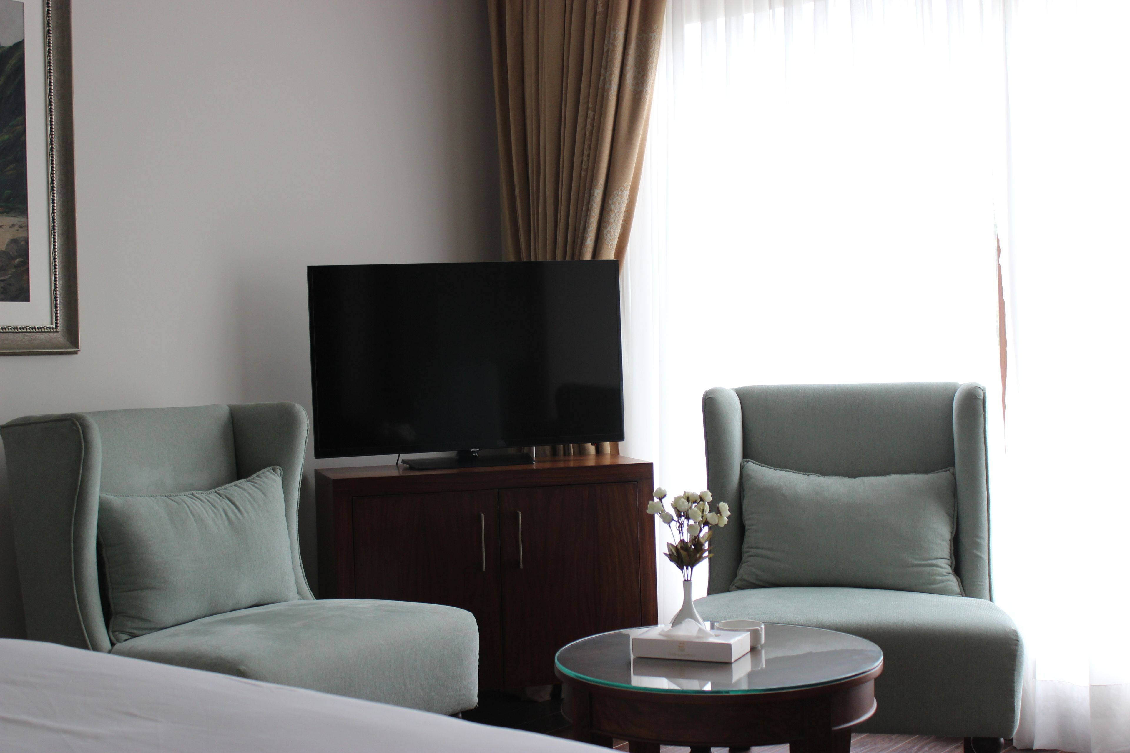 Habitación de hotel con una cama, dos sillones, un televisor sobre un mueble de madera, una mesa de centro de vidrio y un jarrón con flores.