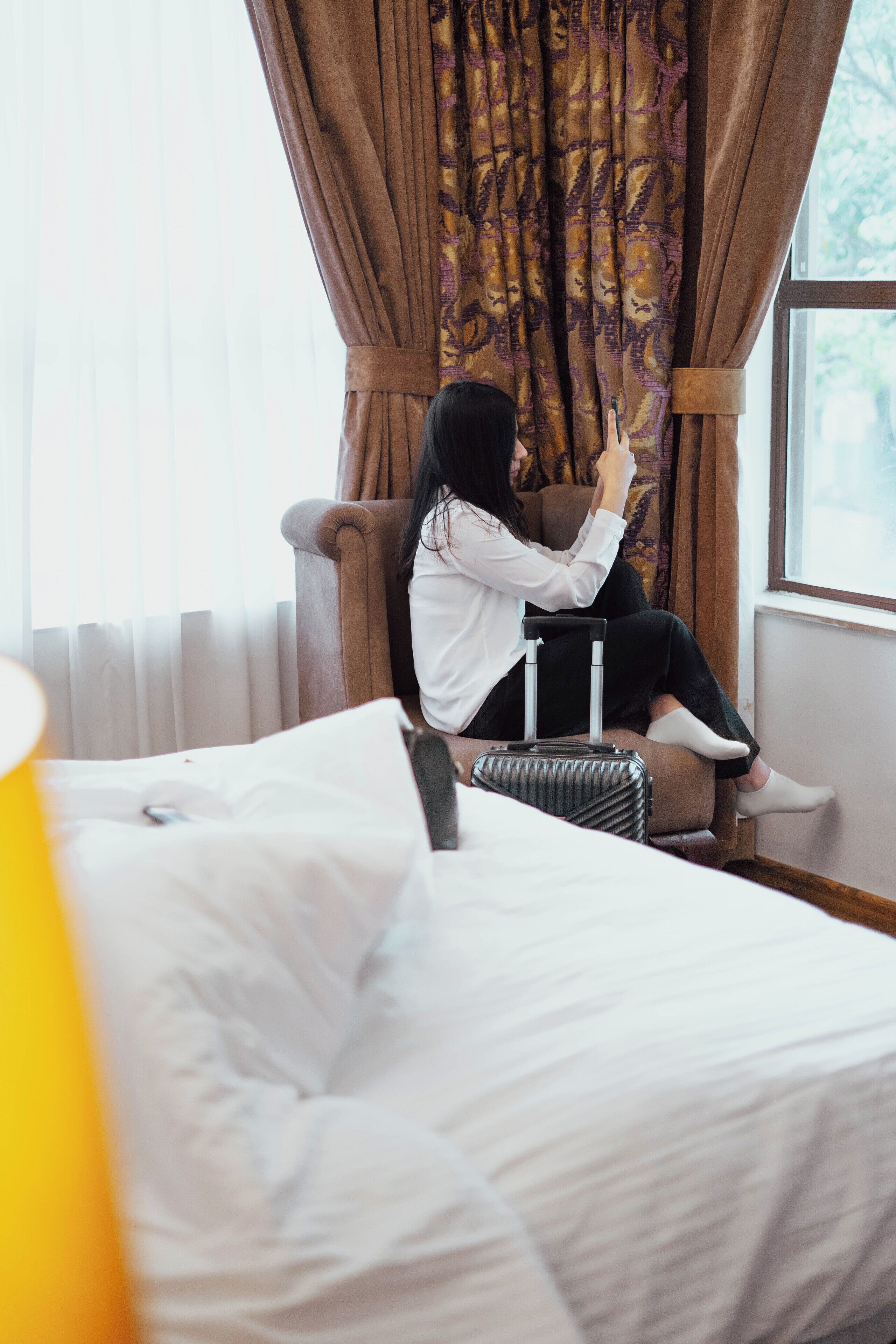 Una persona sentada en una cama en una habitación con ventana y cortinas.