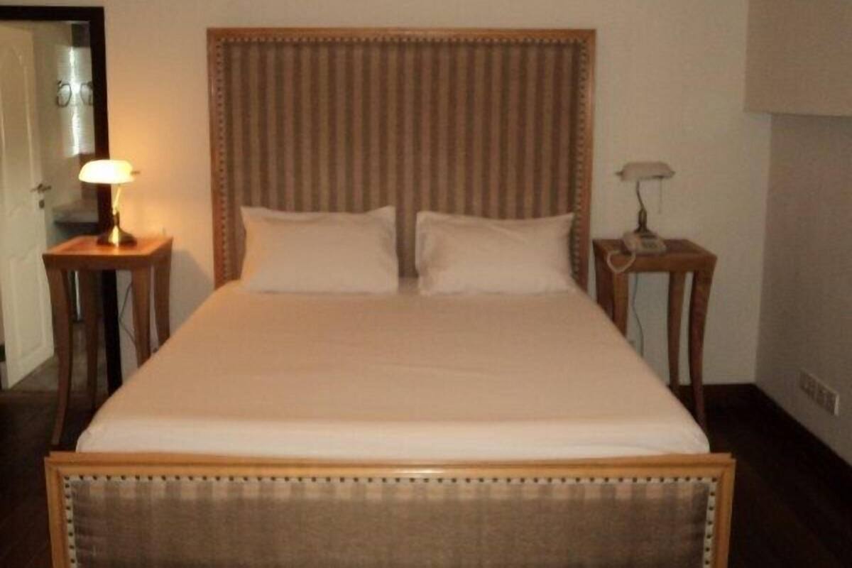 A neatly made bed with a wooden headboard and two pillows, flanked by two bedside tables with lamps, in a room with plain walls.