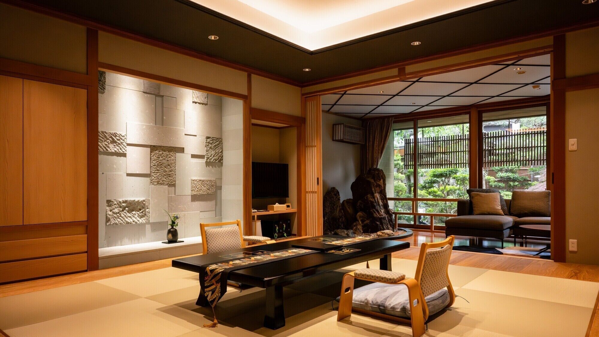 Una habitación japonesa tradicional con suelo de tatami, una mesa baja con camino de mesa negro, una televisión y vistas a un jardín a través de grandes ventanales.