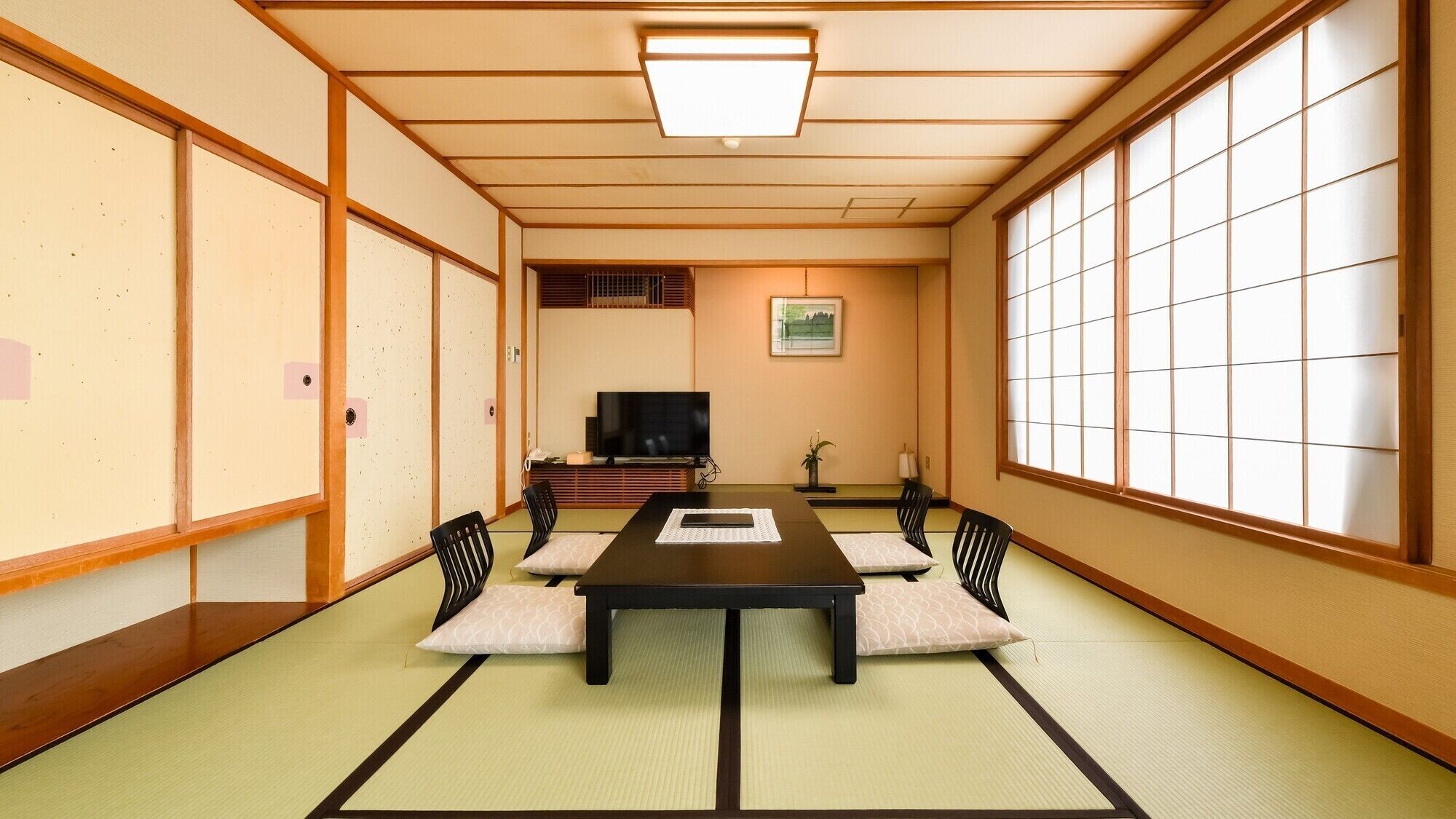Una habitación japonesa tradicional con suelo de tatami, una mesa baja, sillas negras, un televisor y puertas correderas.