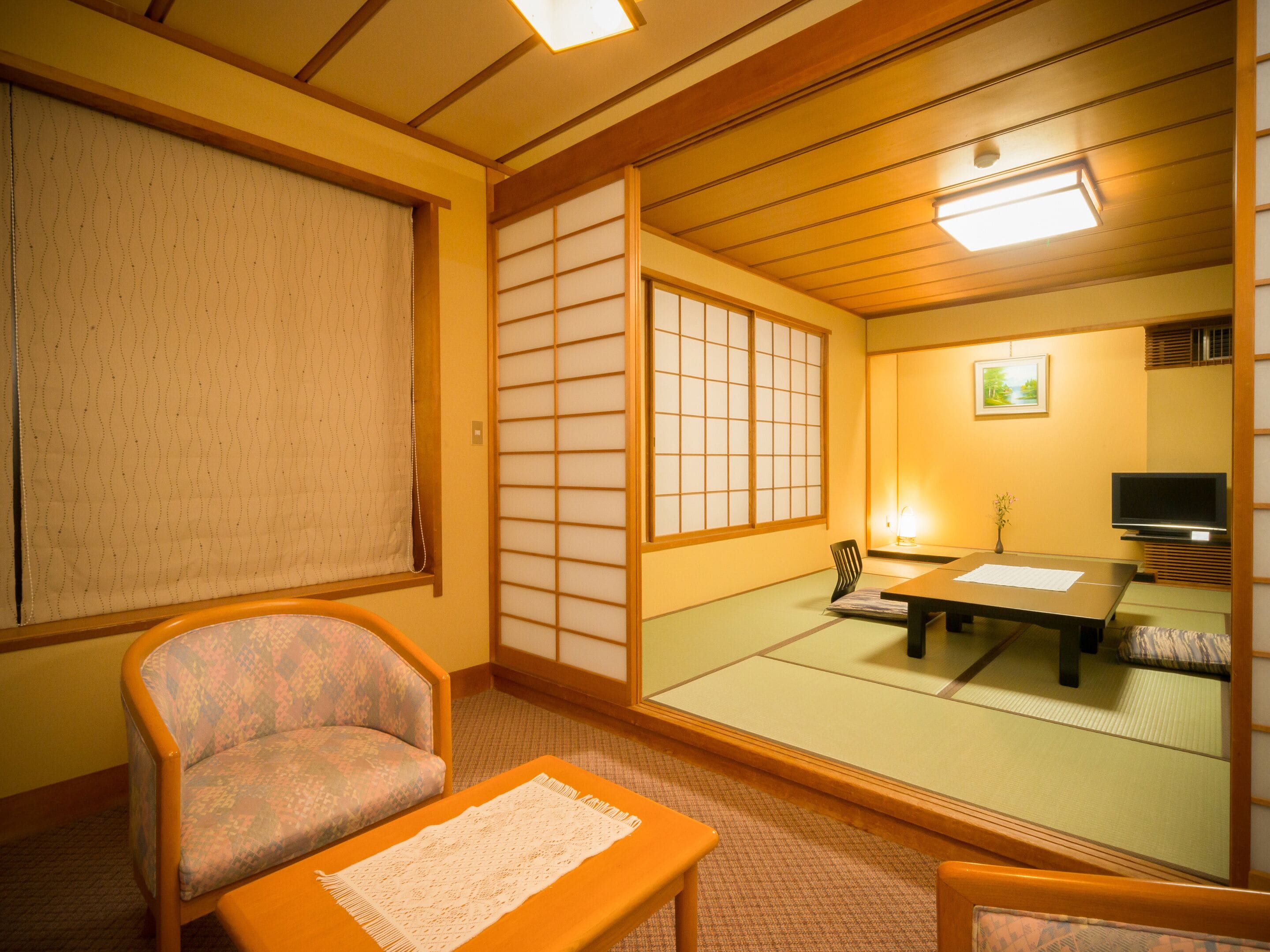 A traditional Japanese room with tatami flooring, a low table, a cushioned chair, a television, and sliding shoji doors.