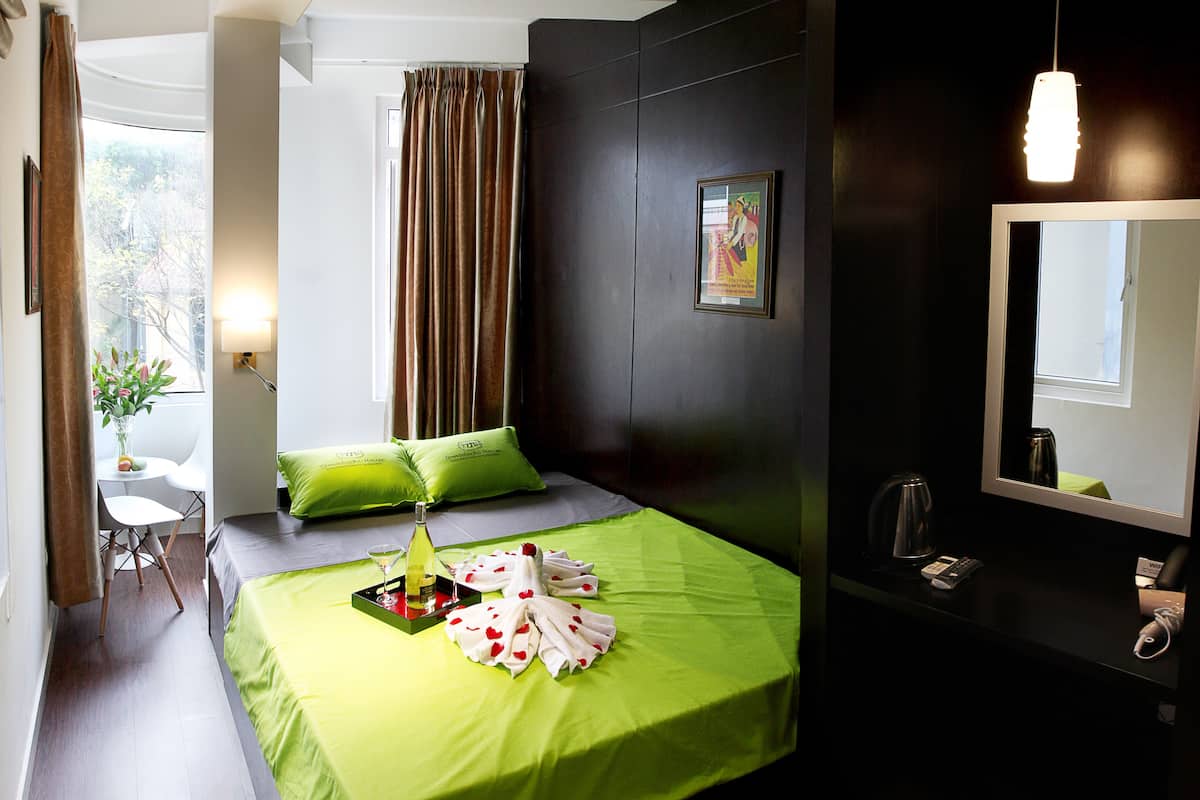 A hotel room with a bed, a desk, a chair, a window with curtains, a wall-mounted mirror, and a bedside table with a kettle and a phone.