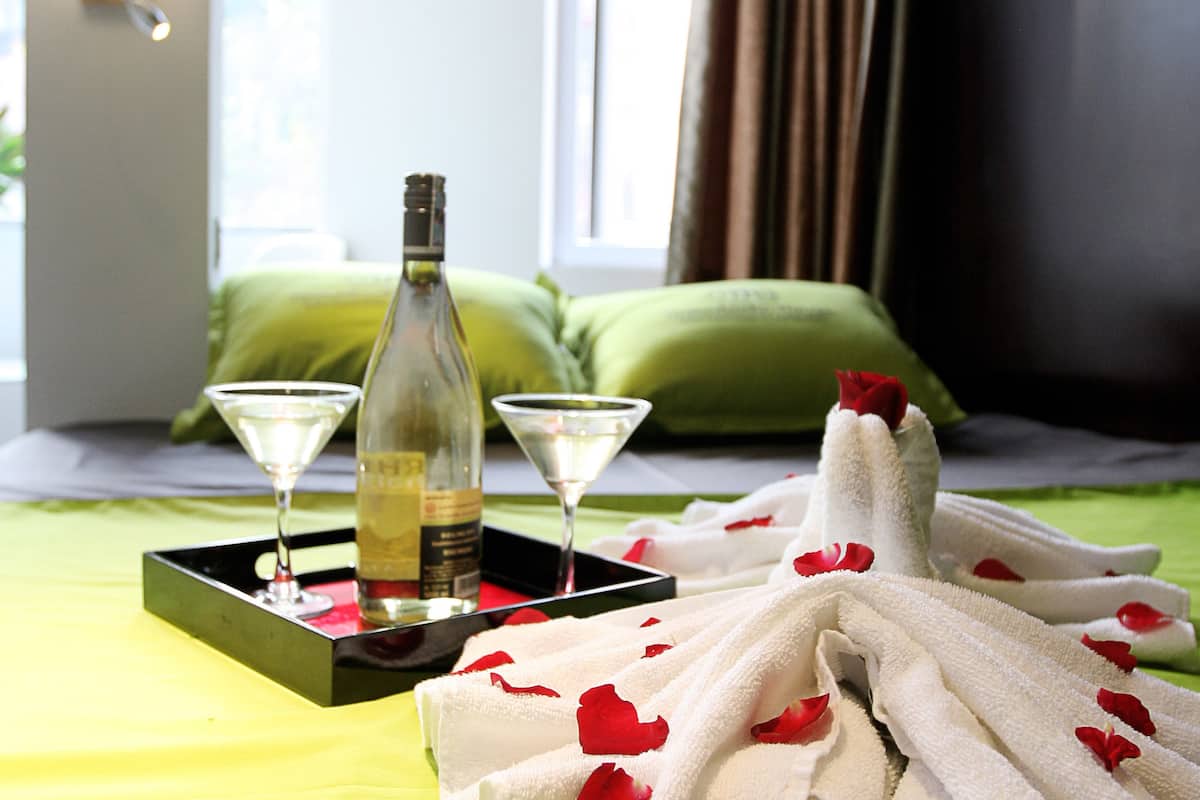 A hotel room with a bed, a bottle of wine, two martini glasses, and a tray with towels and rose petals.