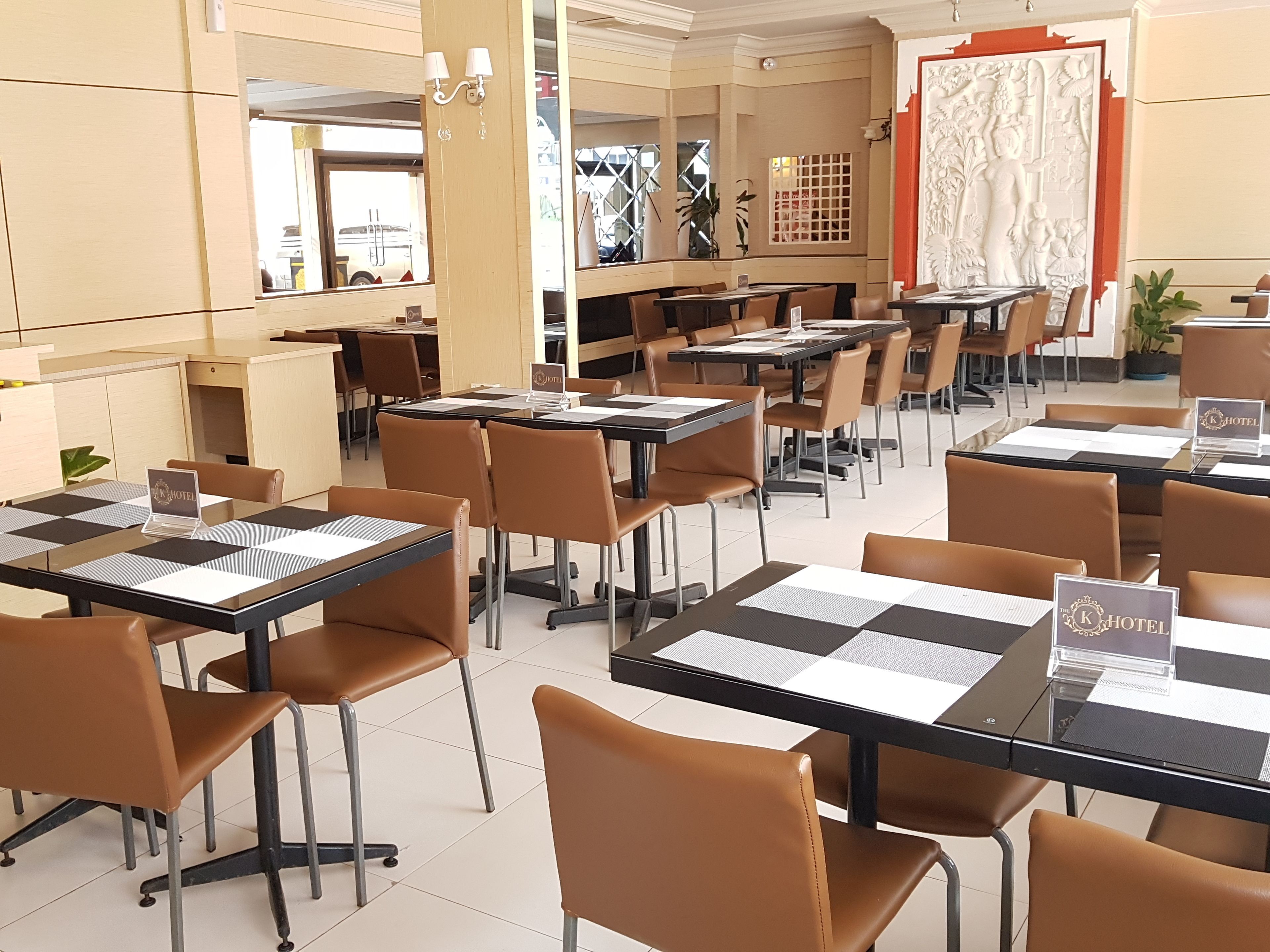 A hotel dining area with multiple tables and chairs, a reception desk, and a decorative wall with a statue.