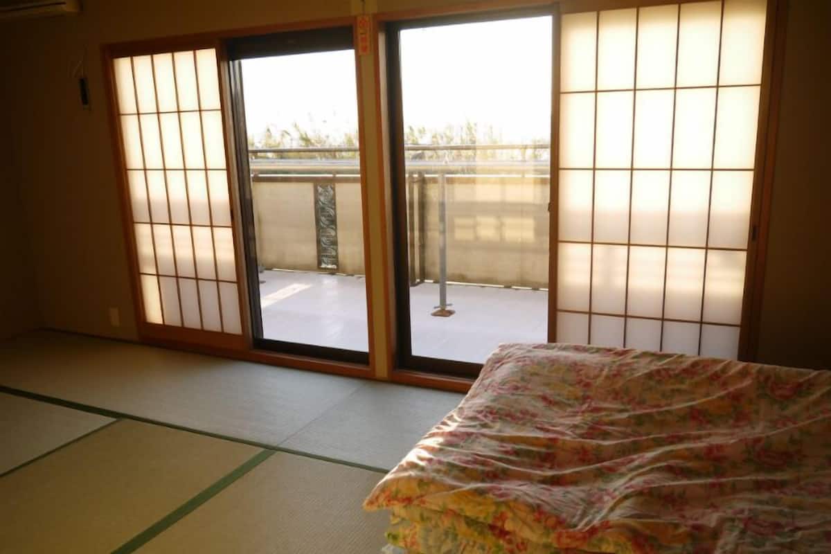 A room with tatami flooring, a bed with a floral bedspread, sliding shoji doors, and a view of a balcony.
