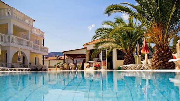 A swimming pool with lounge chairs and umbrellas, a building with balconies, and palm trees. - Zantegarden (Zakynthos)
