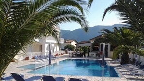 A pool area with lounge chairs, palm trees, and a view of mountains. - Zantegarden (Zakynthos)
