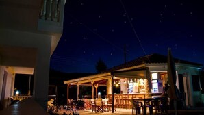 A covered outdoor dining area with tables and chairs, a bar, and a stone building with a tiled roof. - Zantegarden (Zakynthos)
