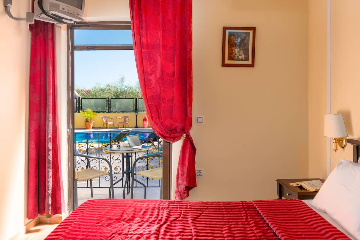 A hotel room with a bed, a window with red curtains, a view of a pool and outdoor seating, and a painting on the wall.