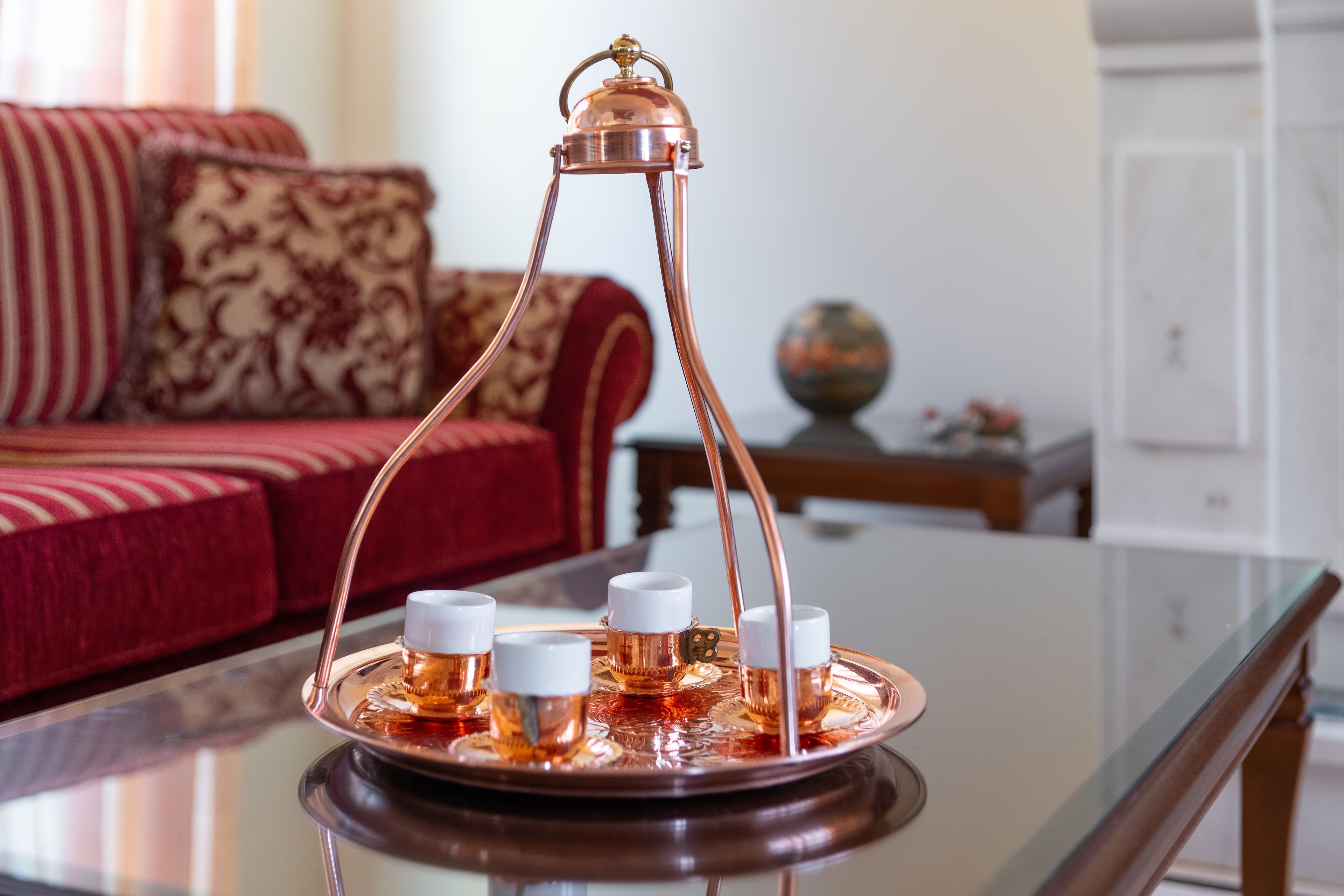 A tray with four glasses of amber liquid, a copper-colored stand, and a white base on a glass table.