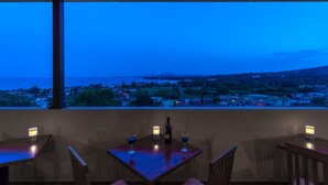 A table with two wine glasses and a bottle, set against a view of a coastal town at dusk. - Hotel Corfu Secret (Corfu)