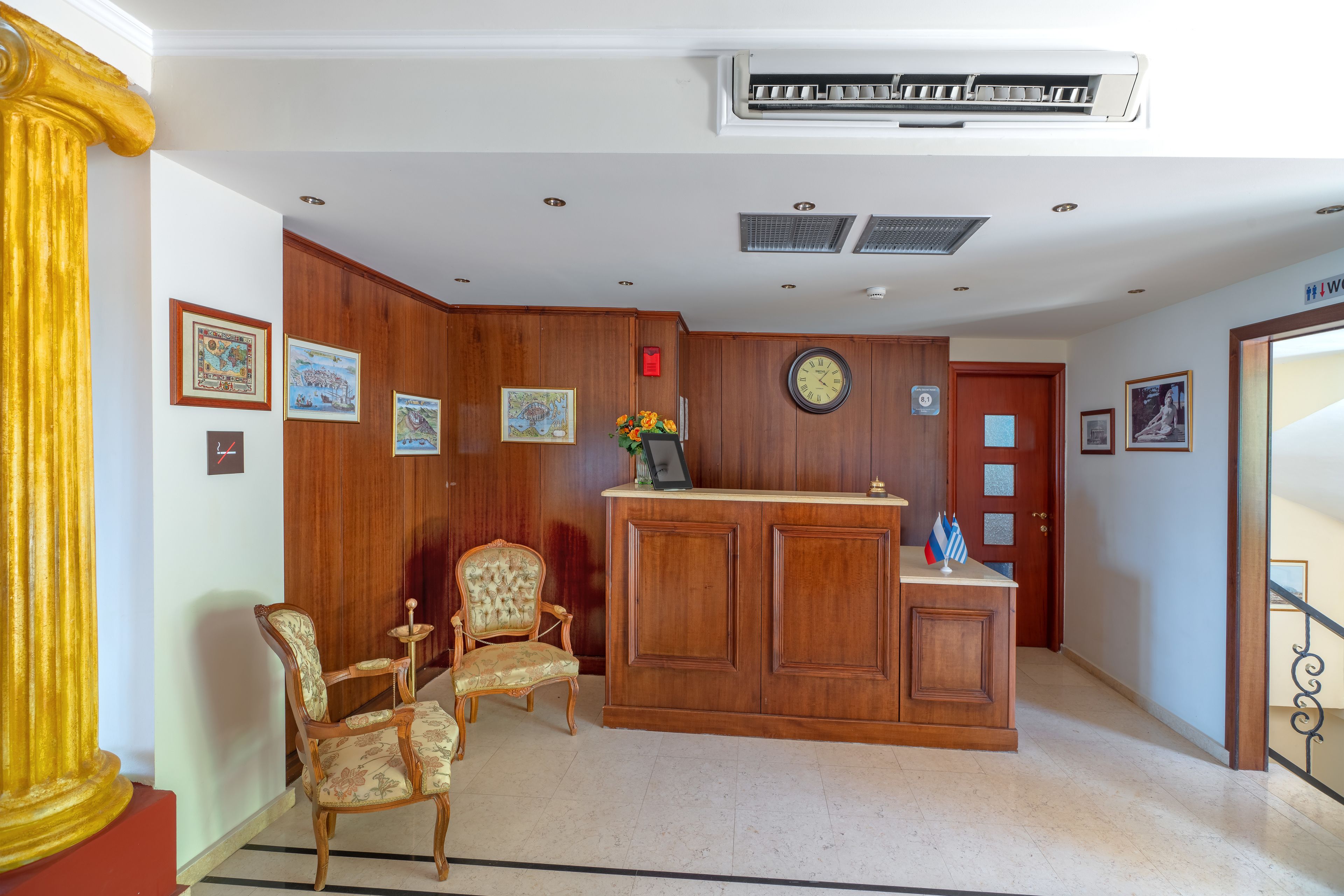 Un área de recepción con un mostrador de madera, dos sillones tapizados, un reloj en la pared y obras de arte enmarcadas.