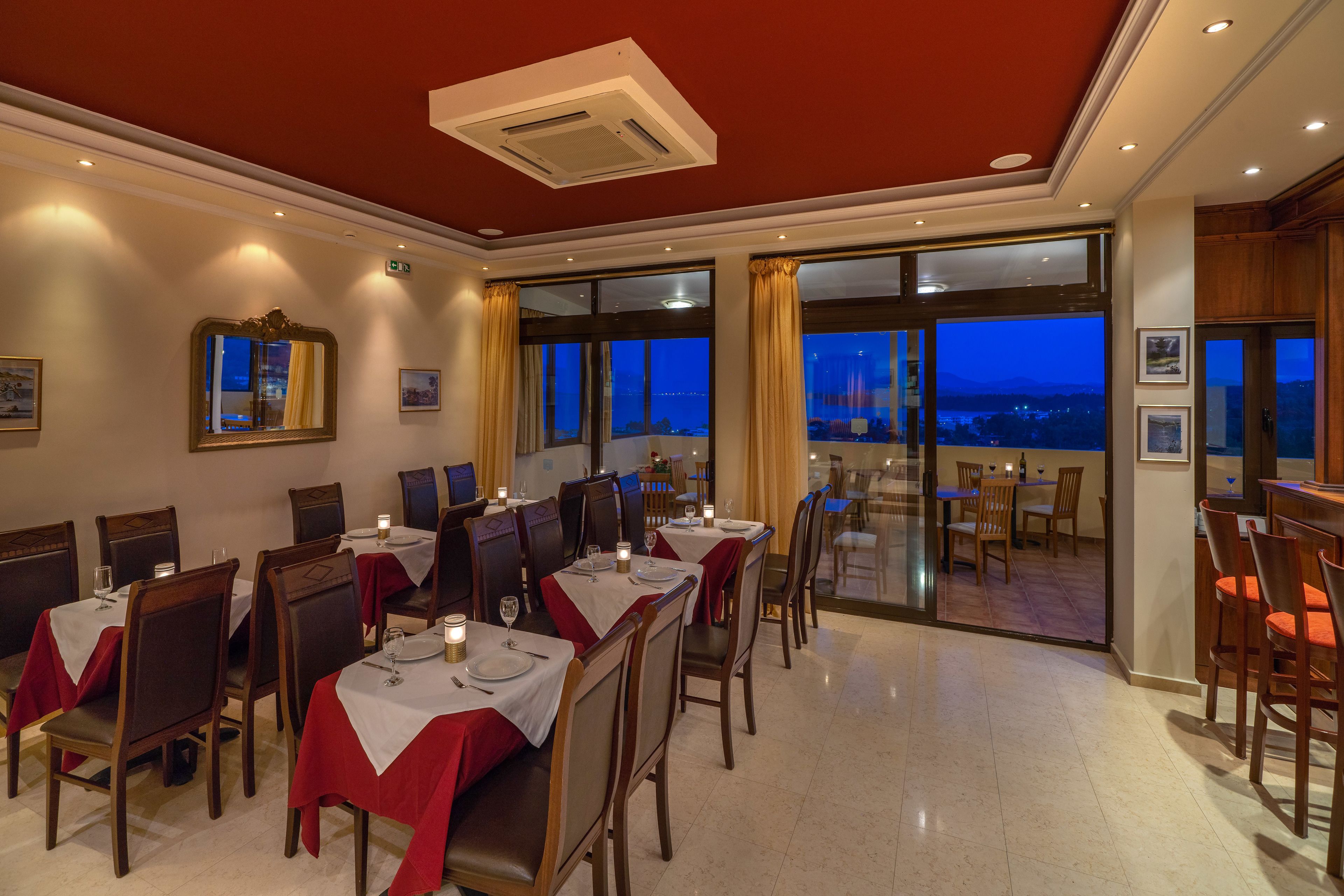 Un comedor con mesas dispuestas para cuatro, ventanas amplias con cortinas y vistas a la ciudad al atardecer.
