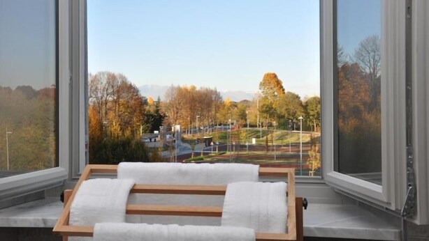 Una habitación con un ventanal grande que da a un parque y una calle. Hay un banco de madera con una toalla blanca sobre él.