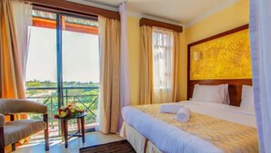 A hotel room with a bed, a chair, a small table with flowers, and a balcony with curtains.