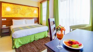 A hotel room with a bed, a table with a fruit platter and a drink, and a chair.