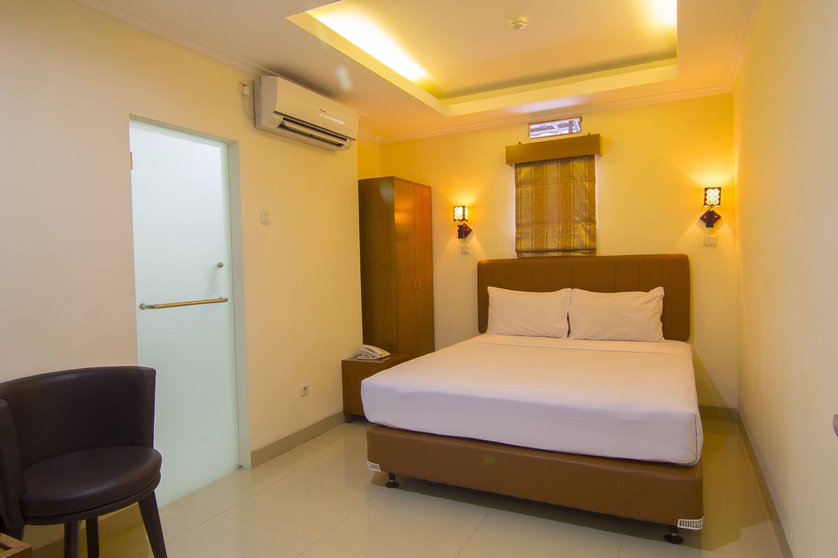 Una habitación de hotel con cama, mesita de noche, silla y unidad de aire acondicionado.