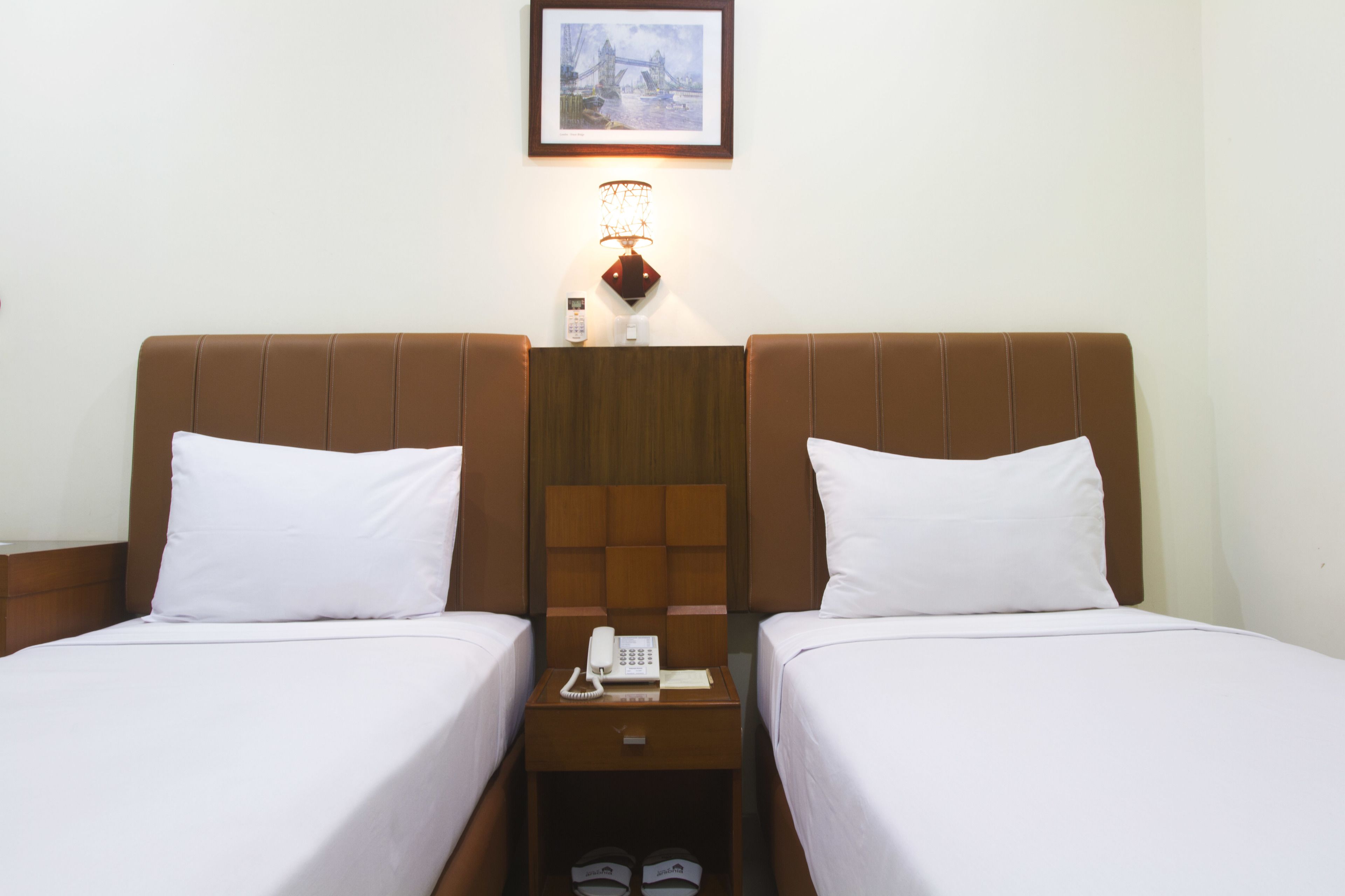 Hotellihuone, jossa on kaksi erillistä sänkyä, yöpöytä, jossa on puhelin ja lamppu, sekä seinällä oleva kehystetty kuva.