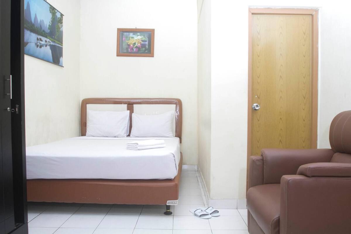A neatly made bed with white linens and pillows, a wooden door, a brown leather chair, and a framed picture on the wall.