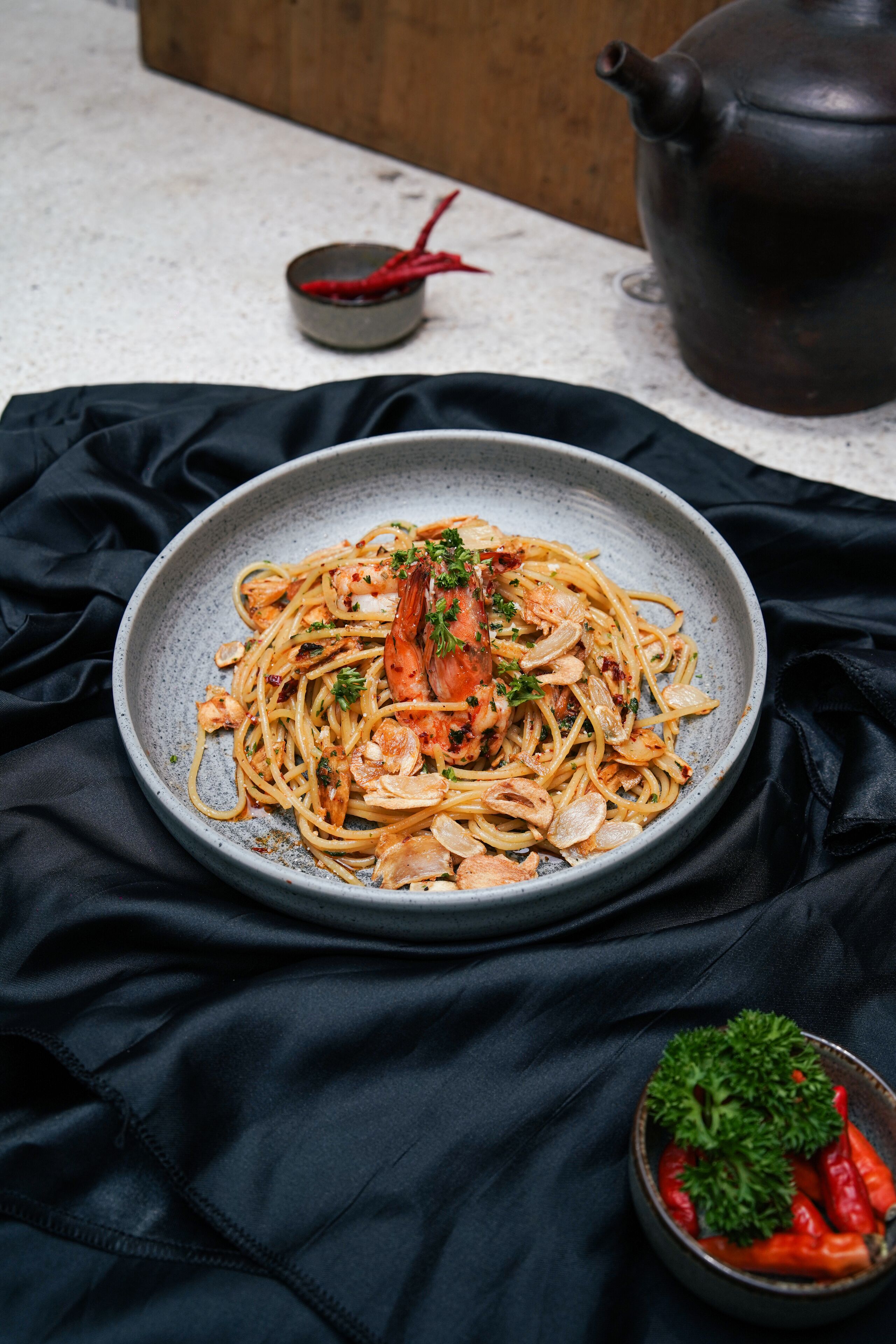A plate of spaghetti with shrimp and herbs, a small bowl of red chili peppers, and a teapot in the background.