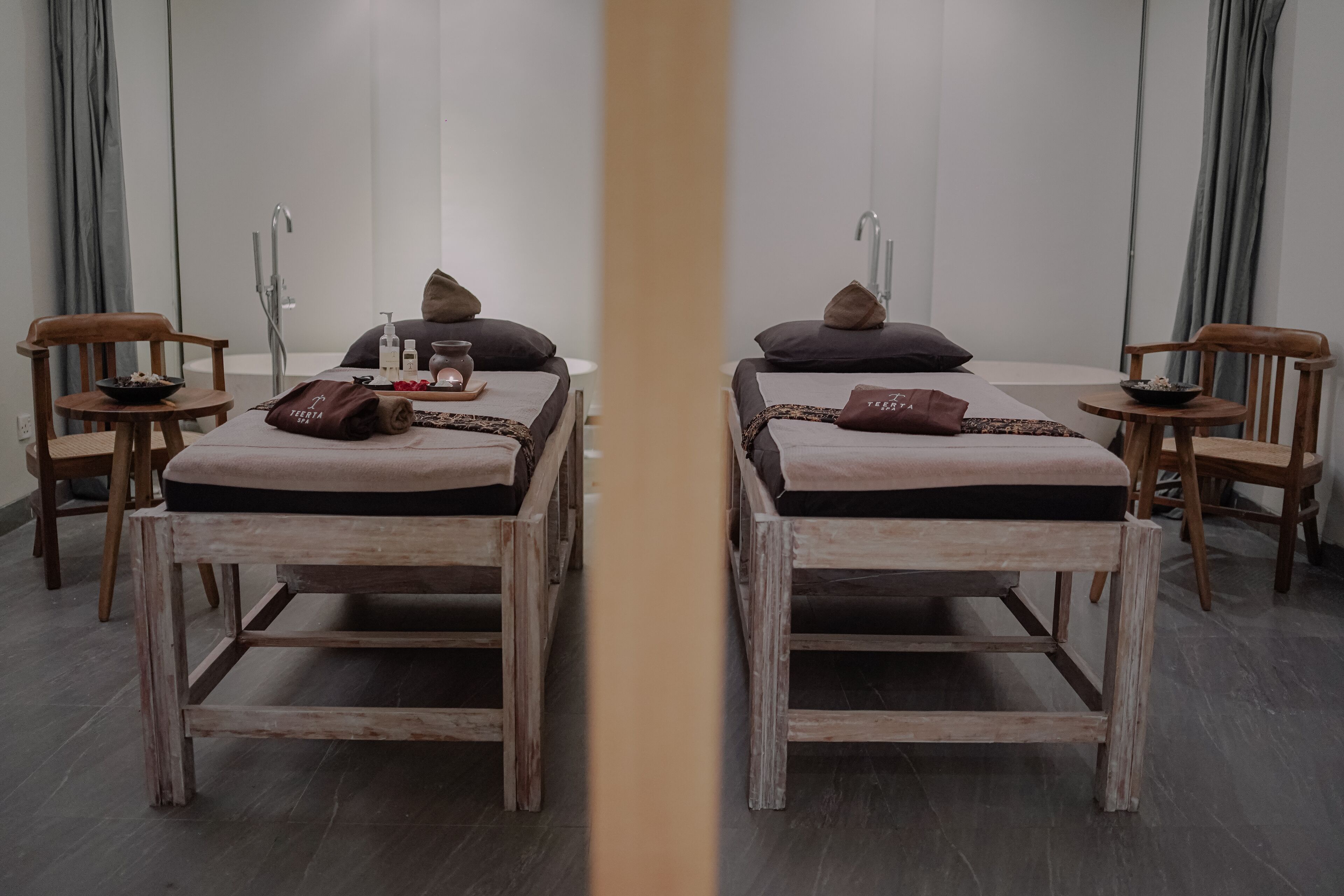 A mirrored room with two wooden beds, each with a pillow and a small table with a bowl and a bottle.
