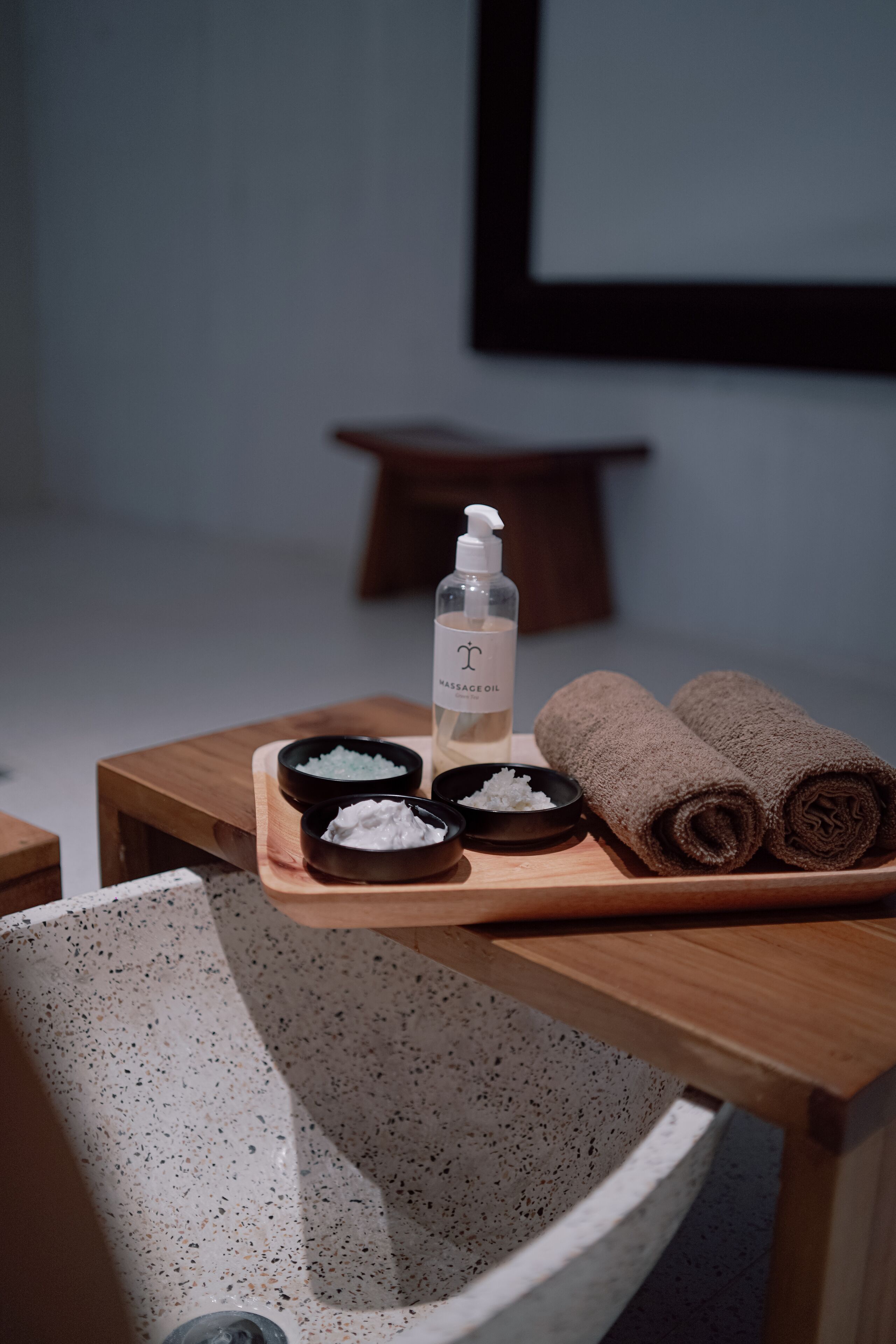 A wooden table with a bottle of massage oil, two bowls of bath salts, and two rolled towels.