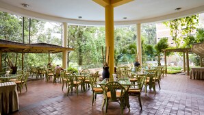 A covered outdoor dining area with green chairs and tables, surrounded by large windows and lush greenery. - Golf Course Hotel (Kampala)