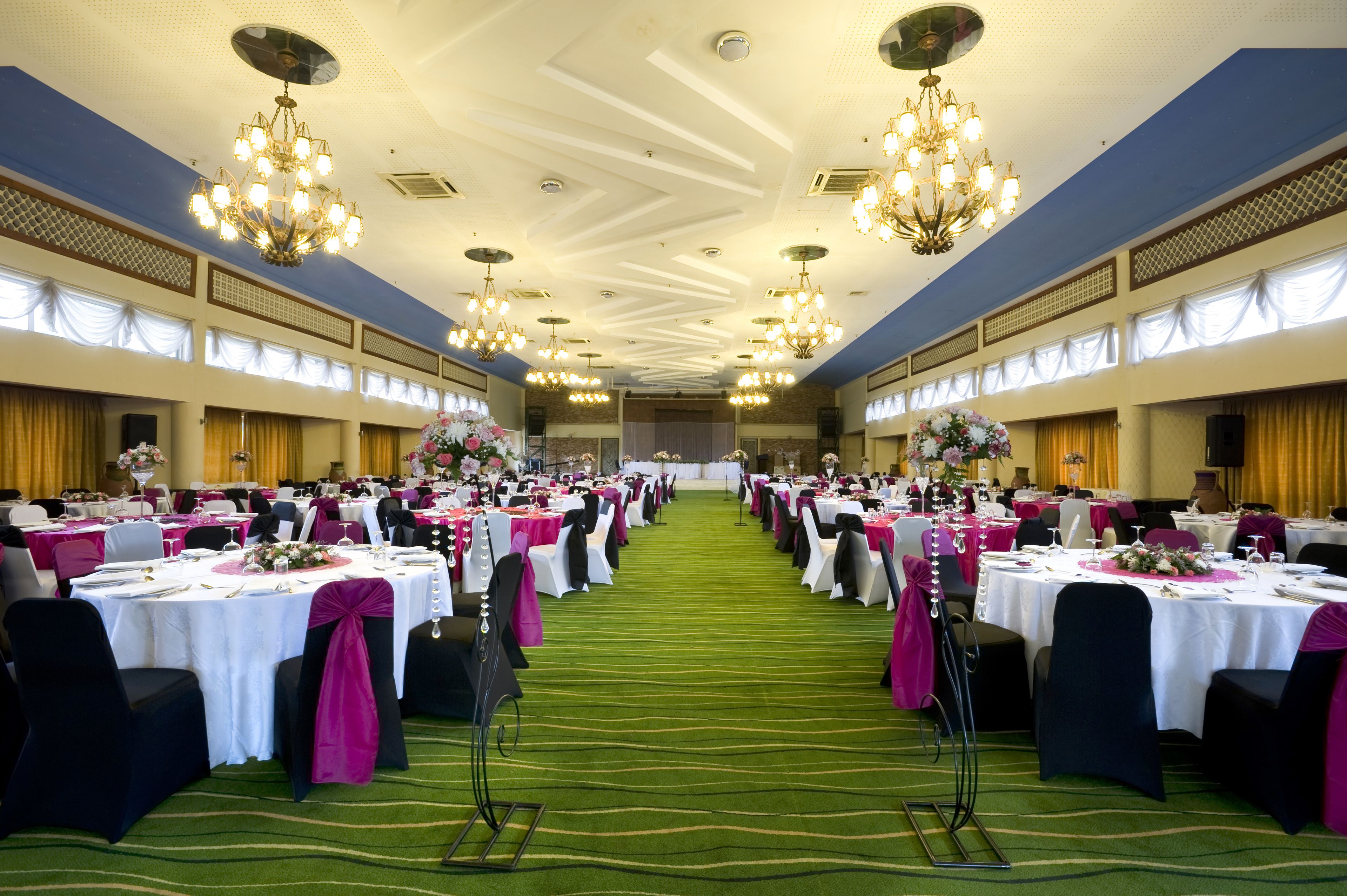 Un salón de banquetes con filas de mesas, cada una con manteles blancos y cubre-sillas rosas, y candelabros colgando del techo.