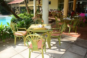 An outdoor dining area with green chairs and yellow tables, surrounded by plants and a pool.