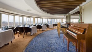 A spacious room with a curved ceiling, large windows offering a city view, a blue patterned carpet, and a grand piano. - Golf Course Hotel (Kampala)