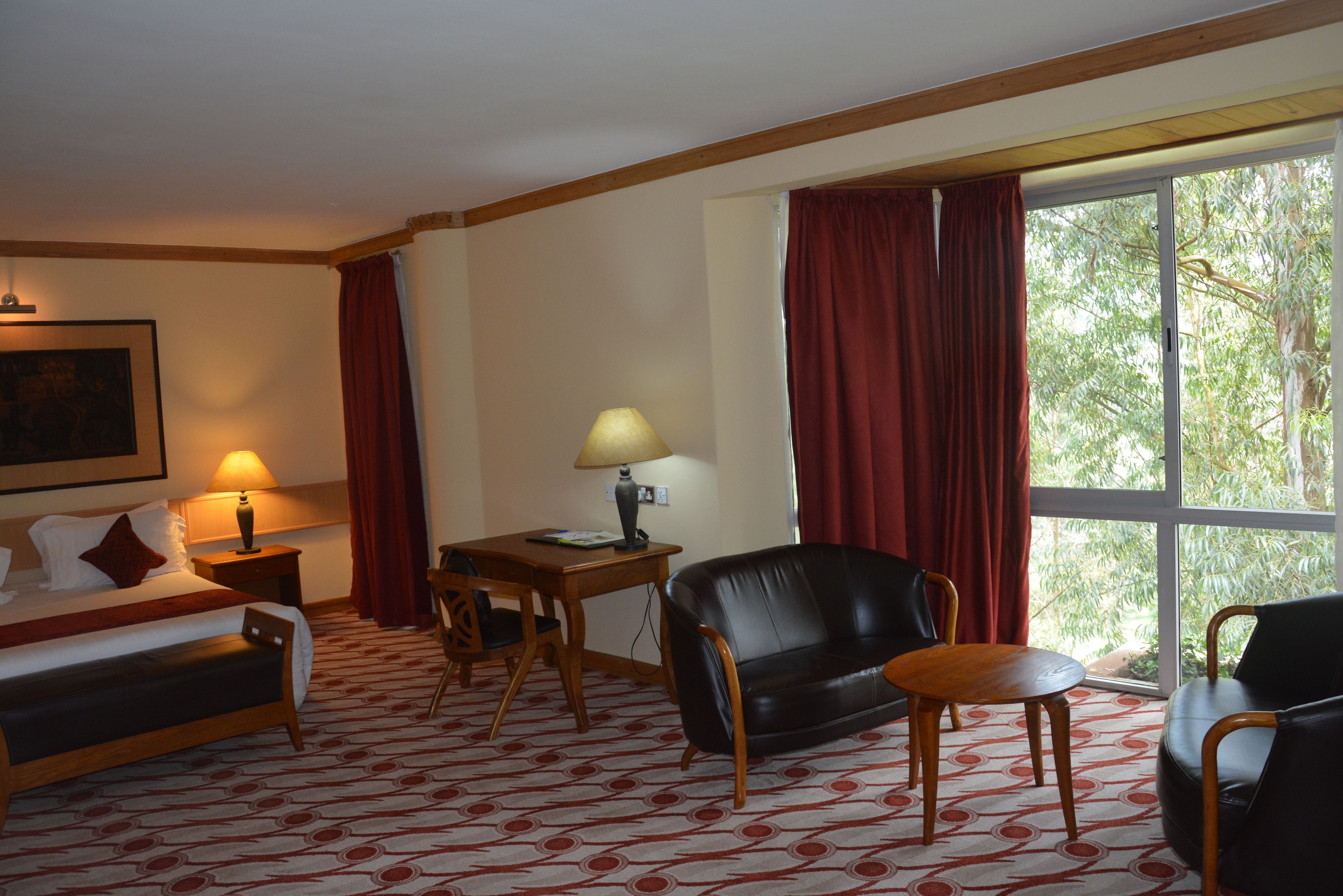 A hotel room with a bed, a desk with a lamp, a leather sofa, and a view of greenery through a large window.