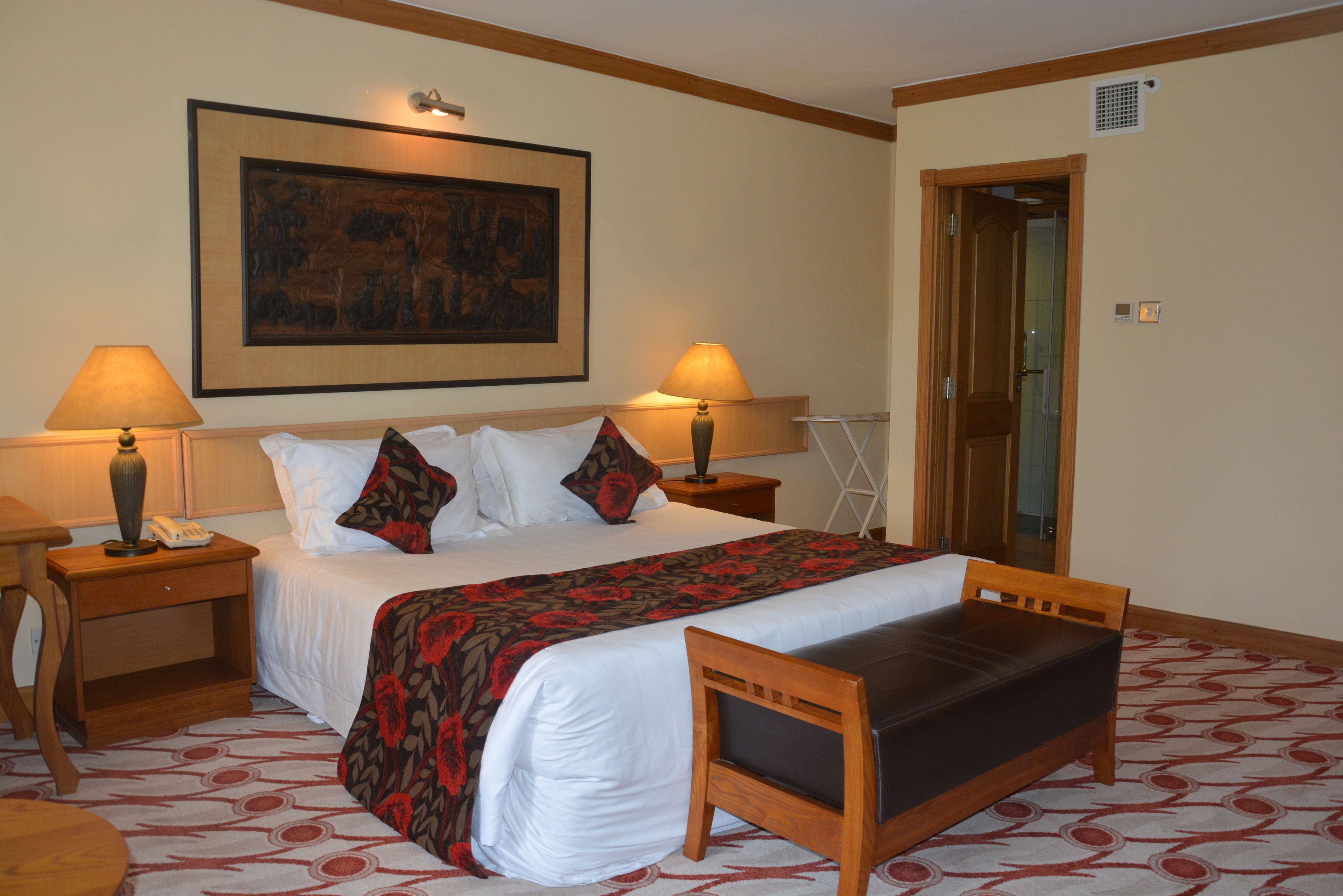 Quarto de hotel com uma cama grande, duas mesas de cabeceira com abajures, um banco de madeira, um tapete estampado e um quadro na parede.