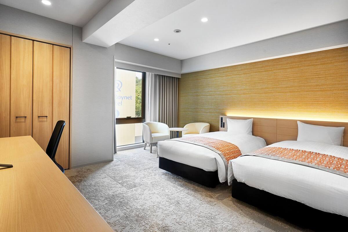 Habitación de hotel con una cama grande, un escritorio y una silla. Tiene una ventana con cortinas, un cabecero de madera y piso alfombrado.