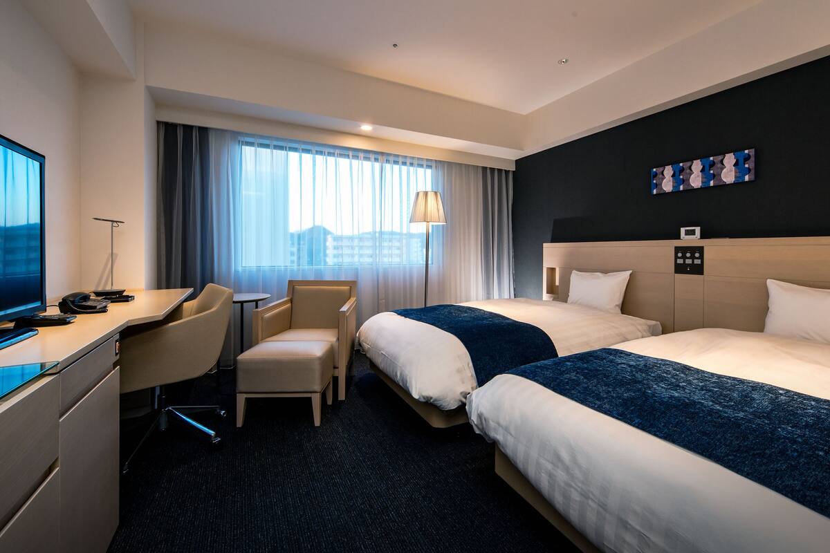 Habitación de hotel con escritorio, dos camas, una silla, televisor y ventana con cortinas.