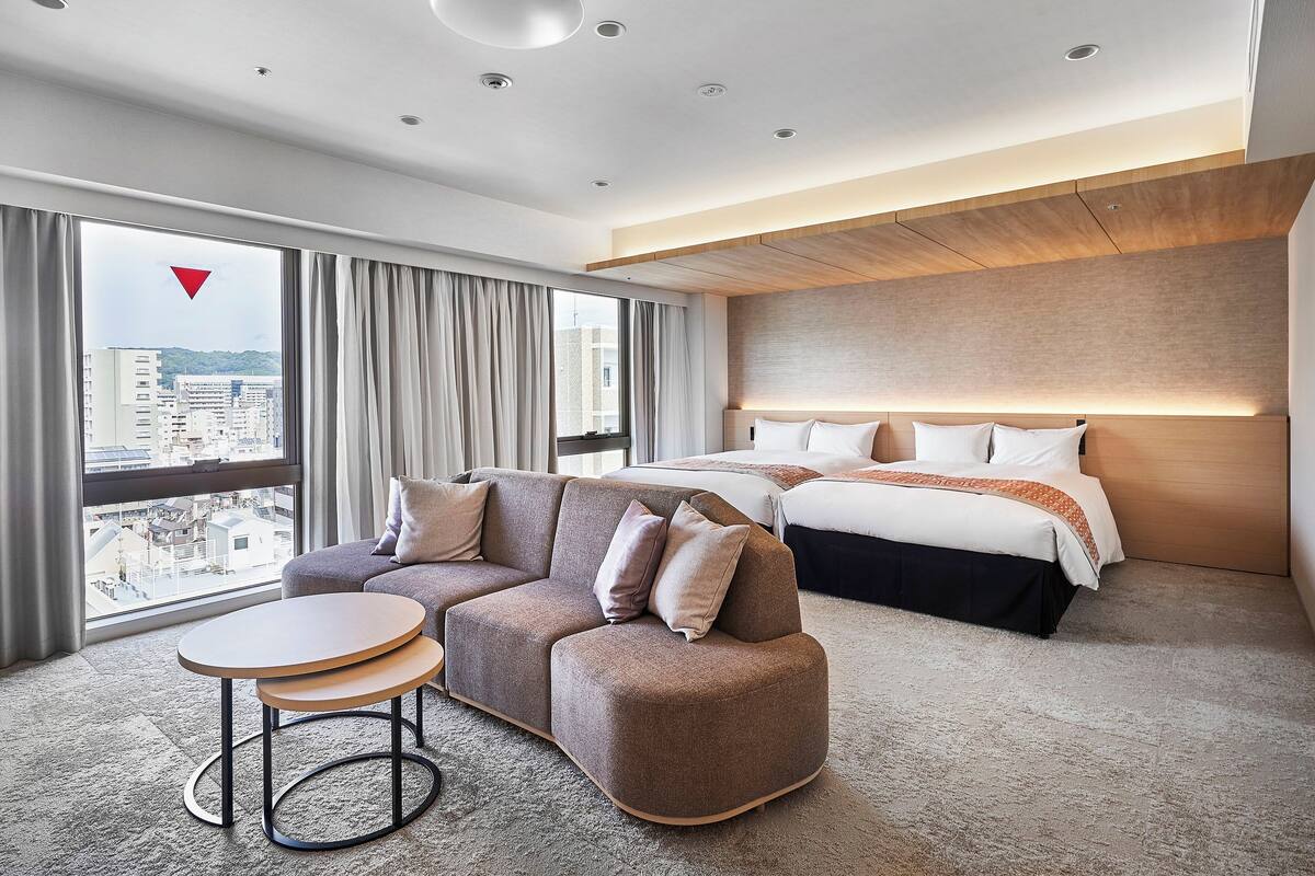 Una habitación de hotel moderna con una cama grande, un sofá, una mesita y vistas a la ciudad a través de amplias ventanas.
