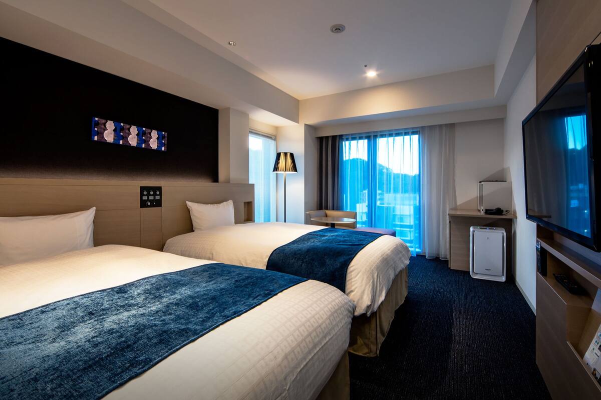 Habitación de hotel con dos camas, televisión, escritorio y ventana con cortinas.