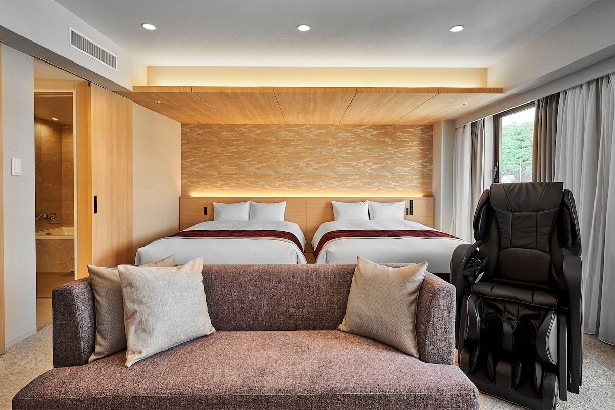 Una habitación de hotel moderna con dos camas, una silla de masaje y un ventanal amplio.