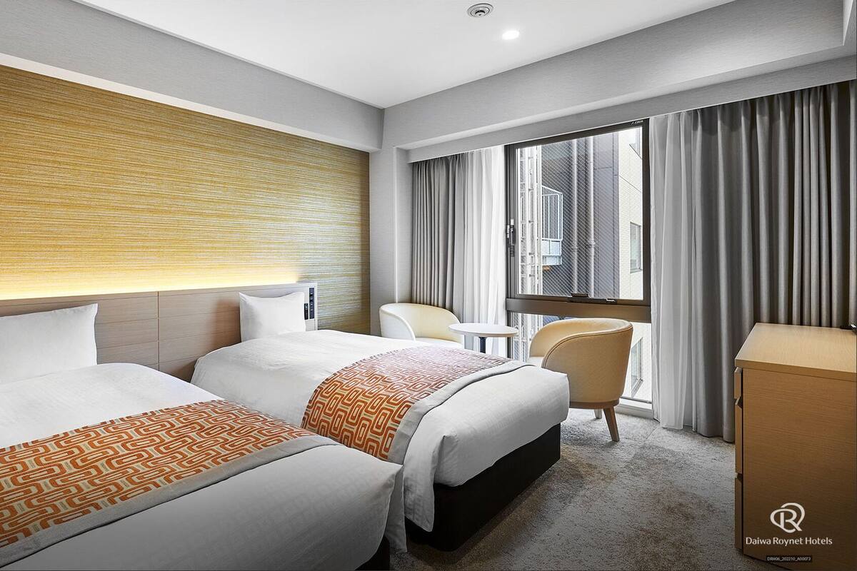 Habitación de hotel con dos camas, una mesita, una silla y vistas de la ciudad a través de amplias ventanas.