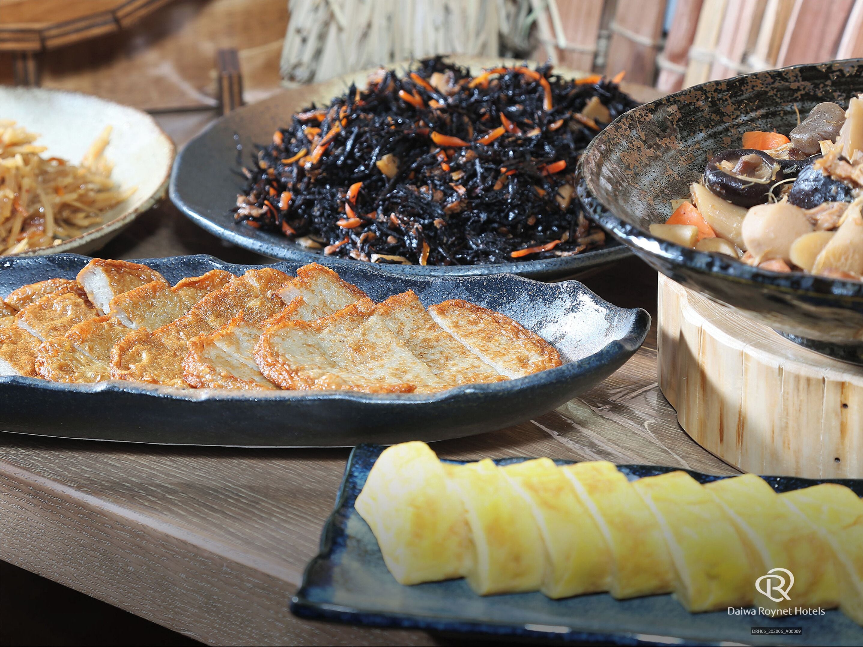 A variety of dishes on a wooden table, including sliced fish, a bowl of blackened vegetables, and a plate of pineapple.